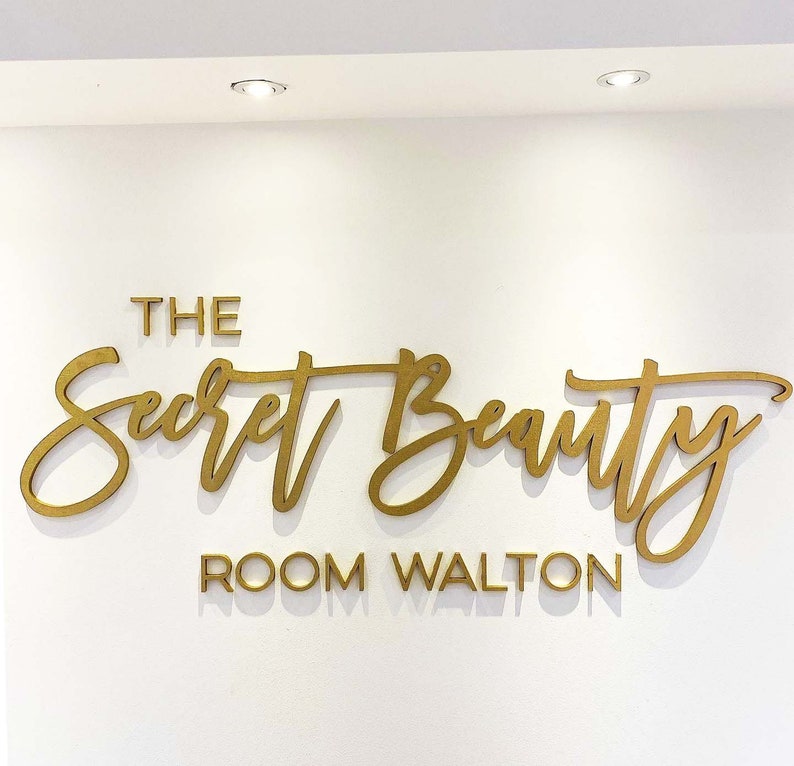 K&ouml;nnte beinhalten: Goldene Beschriftung an einer wei&szlig;en Wand mit dem Text "The Secret Beauty Room Walton".