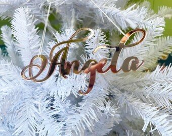 May include: A gold mirror acrylic ornament with the name "Angela" written in a cursive script. The ornament is hanging on a white artificial Christmas tree.