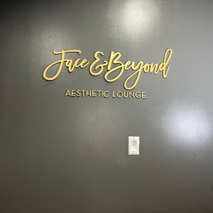K&ouml;nnte beinhalten: Ein goldenes Schild an einer grauen Wand mit der Aufschrift "Face & Beyond Aesthetic Lounge".