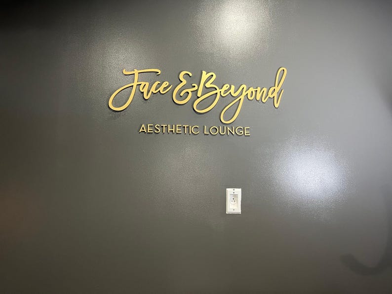 K&ouml;nnte beinhalten: Ein goldenes Schild an einer grauen Wand mit der Aufschrift "Face & Beyond Aesthetic Lounge".
