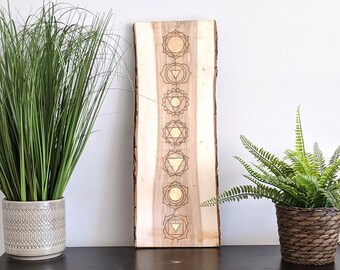 May include: A wooden wall hanging with seven chakra symbols in gold, burned into the wood. The symbols are arranged vertically in a row, with the root chakra at the bottom and the crown chakra at the top. The wood is a light brown color and has a natural grain.