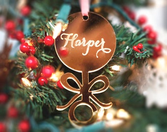 May include: Gold-colored baby rattle ornament with the name "Harper" written in cursive on the round part. The ornament is hanging on a Christmas tree.