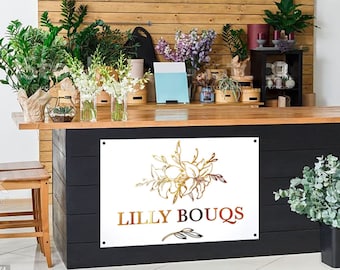 May include: A white sign with a gold floral design and the text "Lilly Bouqs" in gold and red lettering. The sign is mounted on a black wall.