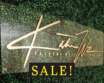 May include: A gold sign with the text "Katerina Mizzi Art Gallery" and a stylized signature. The sign is mounted on a green wall with the word "SALE!" in yellow letters on a black background.