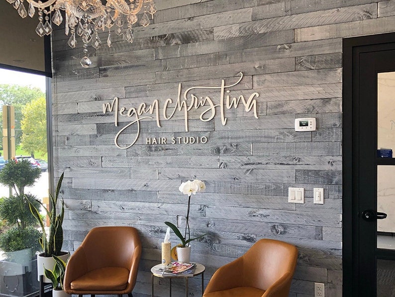 K&ouml;nnte beinhalten: Ein Wartezimmer in einem Friseursalon mit zwei braunen Ledersesseln, einem kleinen Tisch mit einer Zeitschrift und einer wei&szlig;en Orchideenpflanze. Der Name des Salons, "Megan Christina Hair Studio", ist an einer grauen Holzwand angebracht.
