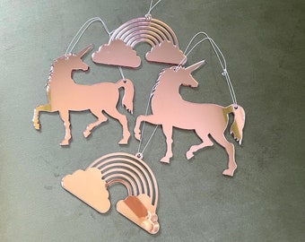 May include: A set of rose gold unicorn and rainbow ornaments. The ornaments are made of mirrored acrylic and feature a unicorn, a rainbow, and a cloud.