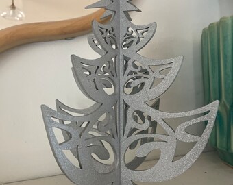 May include: A silver glitter Christmas tree ornament with a star on top and intricate cut-out designs. The tree is made of wood and has a stand.