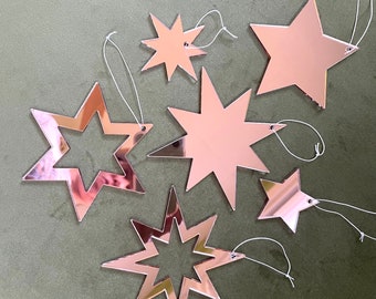 May include: A set of seven rose gold star-shaped ornaments with white string hangers. The stars are various sizes and shapes.