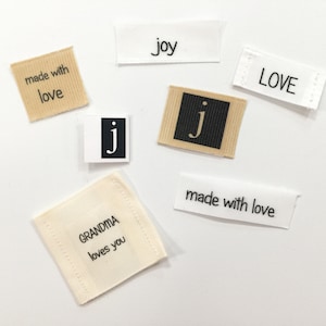 May include: A collection of fabric labels with various messages, including "made with love", "Grandma loves you", "joy", "LOVE", and "j".