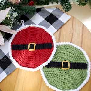 May include: Two handmade crochet pot holders in a Santa Claus design. One is red with a black belt and gold buckle, the other is green with a black belt and gold buckle. Both have a white scalloped edge. The pot holders are on a wooden surface.