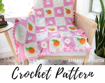 May include: A pink and white crochet blanket with a scalloped edge. The blanket features a repeating pattern of ghosts, pumpkins, and flowers. The blanket is draped over a chair with a wooden frame.