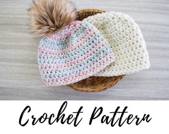 May include: Two crocheted hats, one with a fluffy brown pom-pom, in a wicker basket. The hat with the pom-pom is a multi-colored striped pattern, and the other hat is a solid white.