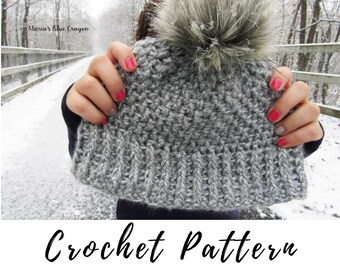 May include: Gray crochet beanie with a fluffy pom pom on top. The beanie is held up in front of a person's face. The text "Crochet Pattern" is at the bottom of the image.