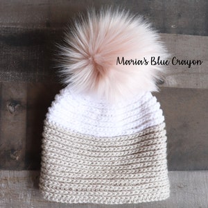 Op de afbeelding: Een beige en witte gebreide beanie met een pluche roze pompon bovenop. De beanie heeft een tweekleurig ontwerp met een witte bovenkant en een beige onderkant.  "Maria's Blue Crayon" staat op de beanie.