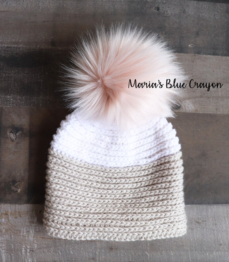Op de afbeelding: Een beige en witte gebreide beanie met een pluche roze pompon bovenop. De beanie heeft een tweekleurig ontwerp met een witte bovenkant en een beige onderkant.  "Maria's Blue Crayon" staat op de beanie.
