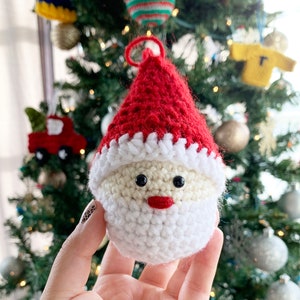 May include: A crocheted Santa Claus ornament with a red hat, white beard, and black eyes. The ornament is hanging on a Christmas tree.