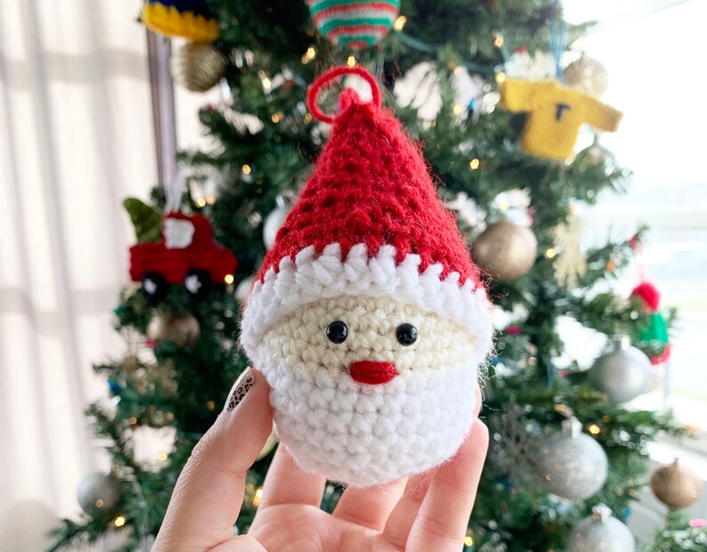 May include: A crocheted Santa Claus ornament with a red hat, white beard, and black eyes. The ornament is hanging on a Christmas tree.