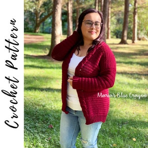 May include: A maroon crocheted cardigan with a cable stitch pattern. The cardigan is open in the front and has long sleeves. The model is wearing the cardigan with a white t-shirt and blue jeans. The text "Maria's Blue Crayon" is visible in the bottom right corner of the image.