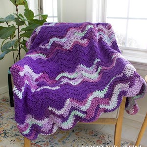 May include: A handmade crochet blanket in shades of purple, pink, and white, draped over a chair. The blanket features a wavy chevron pattern. The text "MARIA'S BLUE CRAYON" is visible in the bottom right corner.