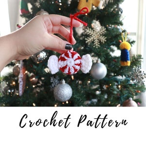 Può includere: Un ornamento a forma di bastoncino di zucchero lavorato a uncinetto con strisce rosse e bianche e anelli bianchi su entrambi i lati. L'ornamento è appeso a uno spago rosso ed è tenuto da una mano davanti a un albero di Natale.