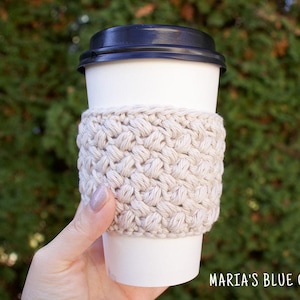 Könnte beinhalten: Ein weißer Kaffeebecher aus Papier mit schwarzem Deckel ist in eine beige, strukturierte, gehäkelte Manschette gehüllt. Die Manschette besteht aus dickem Garn und hat ein gewebtes Muster. Der Text "MARIA'S BLUE CRAYON" befindet sich unten rechts.