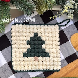 May include: A square crochet potholder featuring a cream-colored background with a dark green pine tree design in the center and a small tan trunk. The text MARIA'S BLUE CRAYON is printed in white above the square, which has a dark green border.