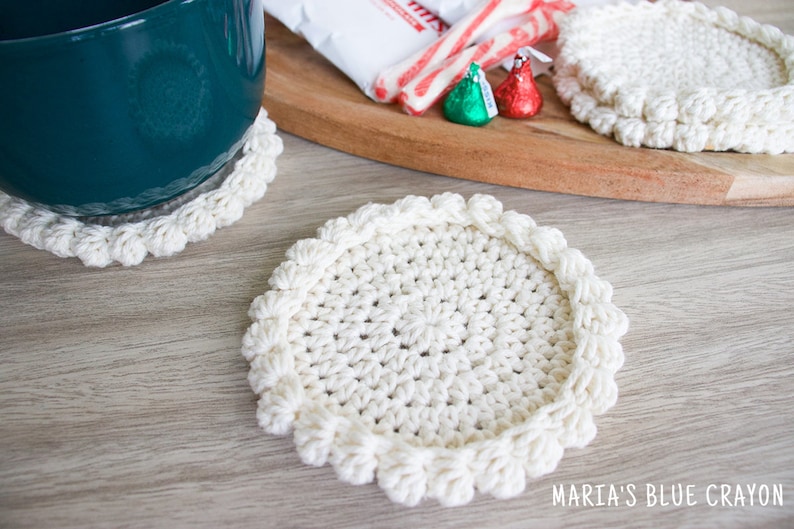 May include: Two white crocheted coasters with a raised edge and a textured center. The coasters are on a light wood surface.