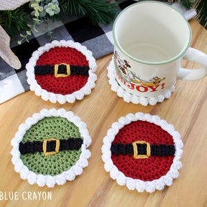 May include: Crocheted coasters in red and green, resembling Santa's belt, with a white scalloped edge. A mug rests on a matching coaster. The coasters are on a wooden surface, with a black and white checkered cloth in the background.