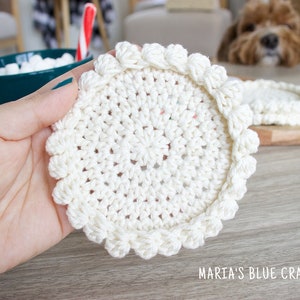 May include: A close-up of a white crocheted coaster with a scalloped edge. The coaster is being held in a hand.