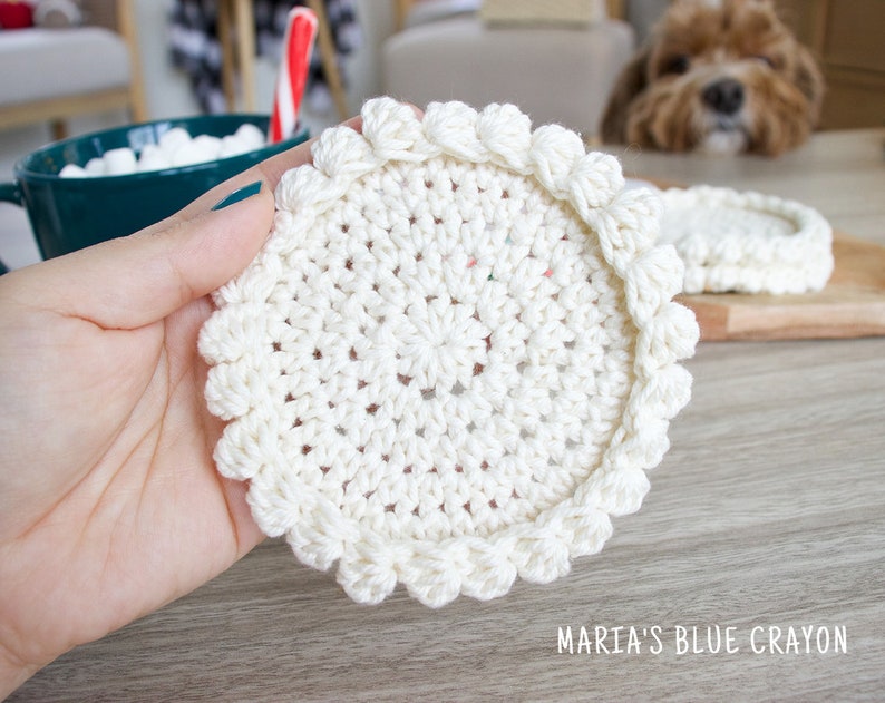 May include: A close-up of a white crocheted coaster with a scalloped edge. The coaster is being held in a hand.