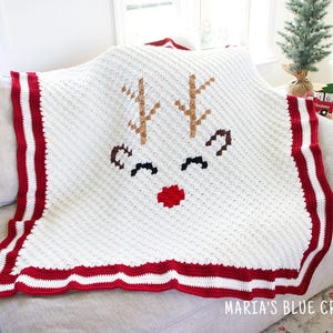 Puede incluir: Una manta blanca de ganchillo con un diseño de cara de reno. La manta tiene un borde a rayas rojo y blanco y presenta cuernos marrones, ojos negros y una nariz roja. El texto "MARIA'S BLUE CRAYON" es visible en la parte inferior.