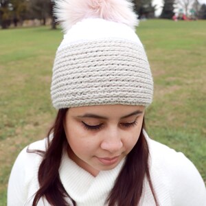 Op de afbeelding: Een witte en beige gebreide muts met een roze nep bont pompon. De muts wordt gedragen door een persoon met lang bruin haar.