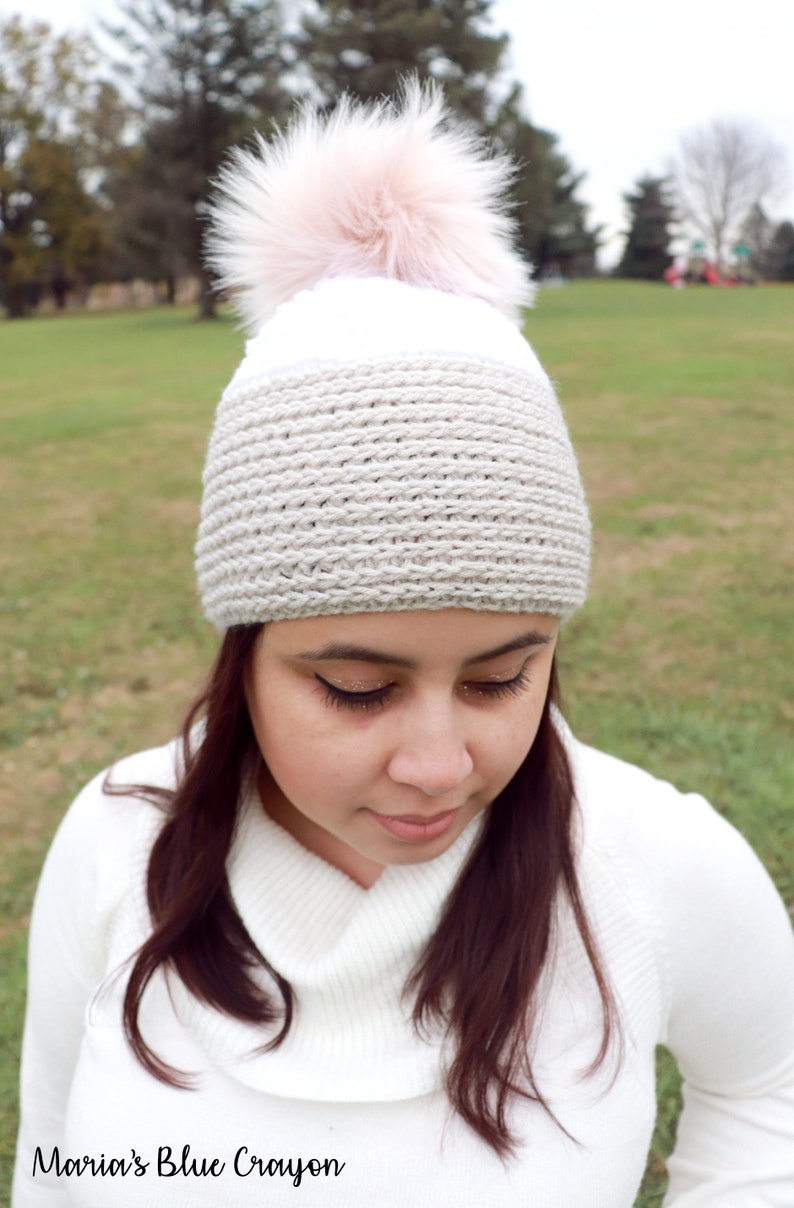 Op de afbeelding: Een witte en beige gebreide muts met een roze nep bont pompon. De muts wordt gedragen door een persoon met lang bruin haar.