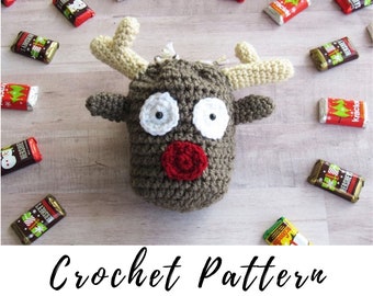 May include: Crochet pattern for a brown reindeer ornament with white eyes, a red nose, and beige antlers. The ornament is surrounded by small chocolate bars.