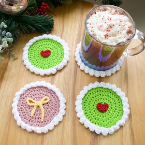 Puede incluir: Una taza de vidrio transparente llena de una bebida caliente y crema batida descansa sobre un posavasos de ganchillo blanco. También se ven tres posavasos de ganchillo adicionales en verde, rosa y blanco. Los posavasos tienen diseños de corazón y lazo.