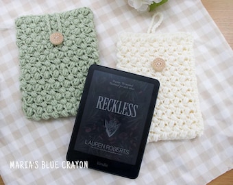 May include: Two handmade crochet Kindle covers in sage green and cream, each with a wooden button closure. A Kindle e-reader with the book title "Reckless" is placed between the covers. The covers are on a checkered fabric.