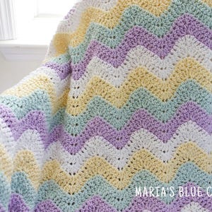 May include: A crocheted chevron blanket in shades of purple, green, yellow, and white. The blanket is made with a soft yarn and has a delicate texture.