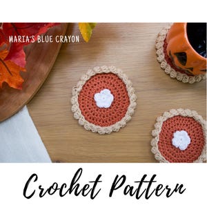 Peut inclure: Des dessous de verre au crochet en forme de tarte. Les dessous de verre sont de couleur rouille avec une croûte beige et une fleur blanche au centre. Le texte "Maria's Blue Crayon" et "Crochet Pattern" sont également visibles.