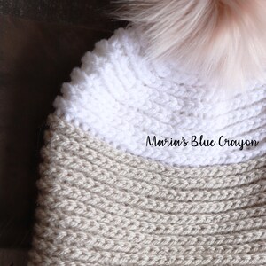 Op de afbeelding: Een beige en witte gebreide muts met een pluche roze pompon bovenop. De muts heeft een geribbelde textuur en de tekst "Maria's Blue Crayon" is zichtbaar op de muts.