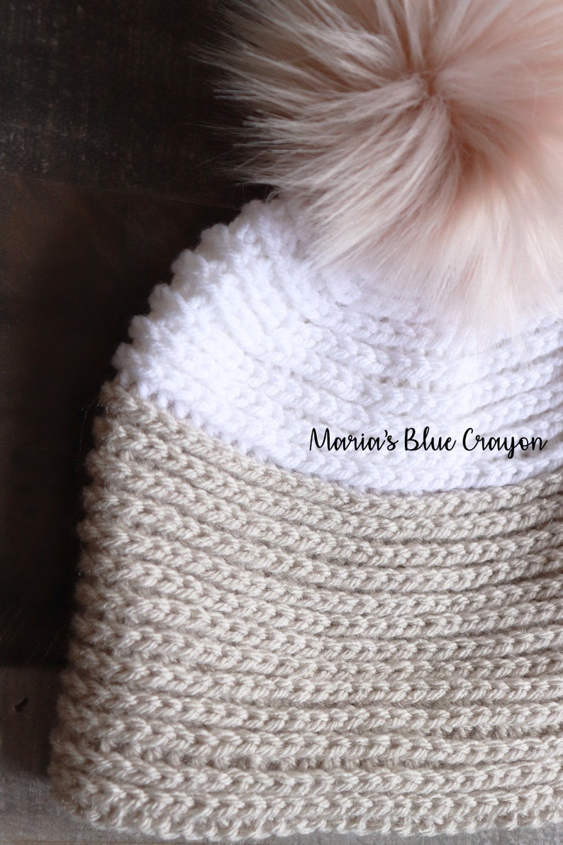 Op de afbeelding: Een beige en witte gebreide muts met een pluche roze pompon bovenop. De muts heeft een geribbelde textuur en de tekst "Maria's Blue Crayon" is zichtbaar op de muts.