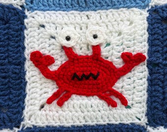 May include: A crocheted square featuring a red crab with black eyes and a black mouth, set against a white background. The square is framed by blue and white crochet. The text "MARIA'S BLUE CRAYON" is in the bottom right corner.