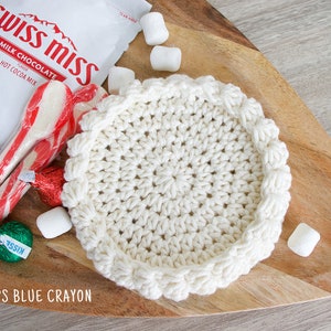 May include: A white crocheted coaster with a scalloped edge sits on a wooden surface.  There are candy canes, marshmallows, and a chocolate kiss on the surface.
