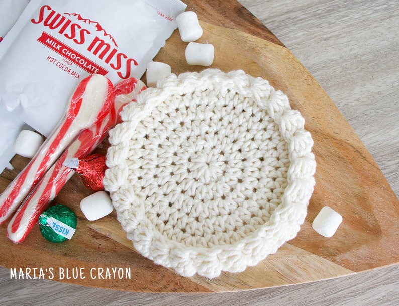 May include: A white crocheted coaster with a scalloped edge sits on a wooden surface.  There are candy canes, marshmallows, and a chocolate kiss on the surface.