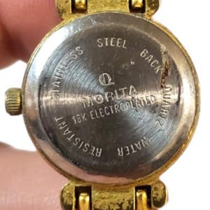 May include: Close-up of a vintage watch with a pearl bracelet. The watch face is gold-toned with a stainless steel back, displaying the text "MORITA" and "18K ELECTROPLATED". The bracelet features two strands of cream-colored faux pearls.