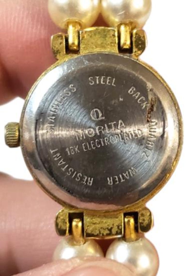 May include: Close-up of a vintage watch with a pearl bracelet. The watch face is gold-toned with a stainless steel back, displaying the text "MORITA" and "18K ELECTROPLATED". The bracelet features two strands of cream-colored faux pearls.