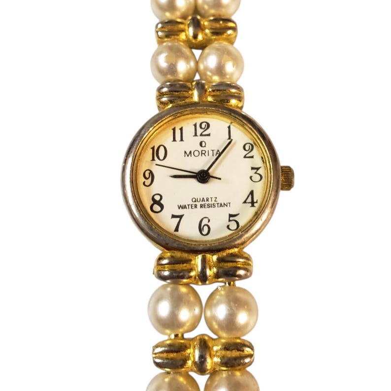May include: A gold-toned Morita quartz watch with a white face, black numerals, and hands. The watchband is made of faux pearls. The watch face reads "QUARTZ WATER RESISTANT".