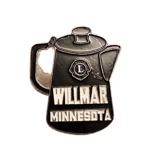 May include: A black and silver pin in the shape of a coffee pot. The pin features the text "WILLMAR MINNESOTA" in white lettering. The Lions Club logo is visible on the coffee pot. The pin is likely a souvenir or collectible item.