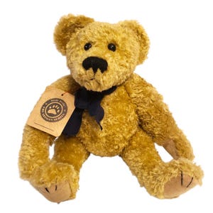 May include: A golden-brown teddy bear with a black nose and a black ribbon around its neck. The bear has a tag attached with a paw print design. The plush toy is sitting with its legs spread.