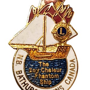 May include: A gold-toned metal lapel pin featuring a white sailing ship with red sails and a Lions Club emblem. The circular base contains blue textured water and the text: The Bay Chaleur Phantom Ship, N.B. BATHURST LIONS CANADA.