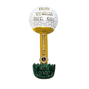 May include: A white golf ball pin with gold lettering reading DEAN ST EINER DIST GOV. The pin features a gold vertical post with a small circular emblem containing an L, and a green base with the text 5M-2 84-85 embossed in gold lettering.